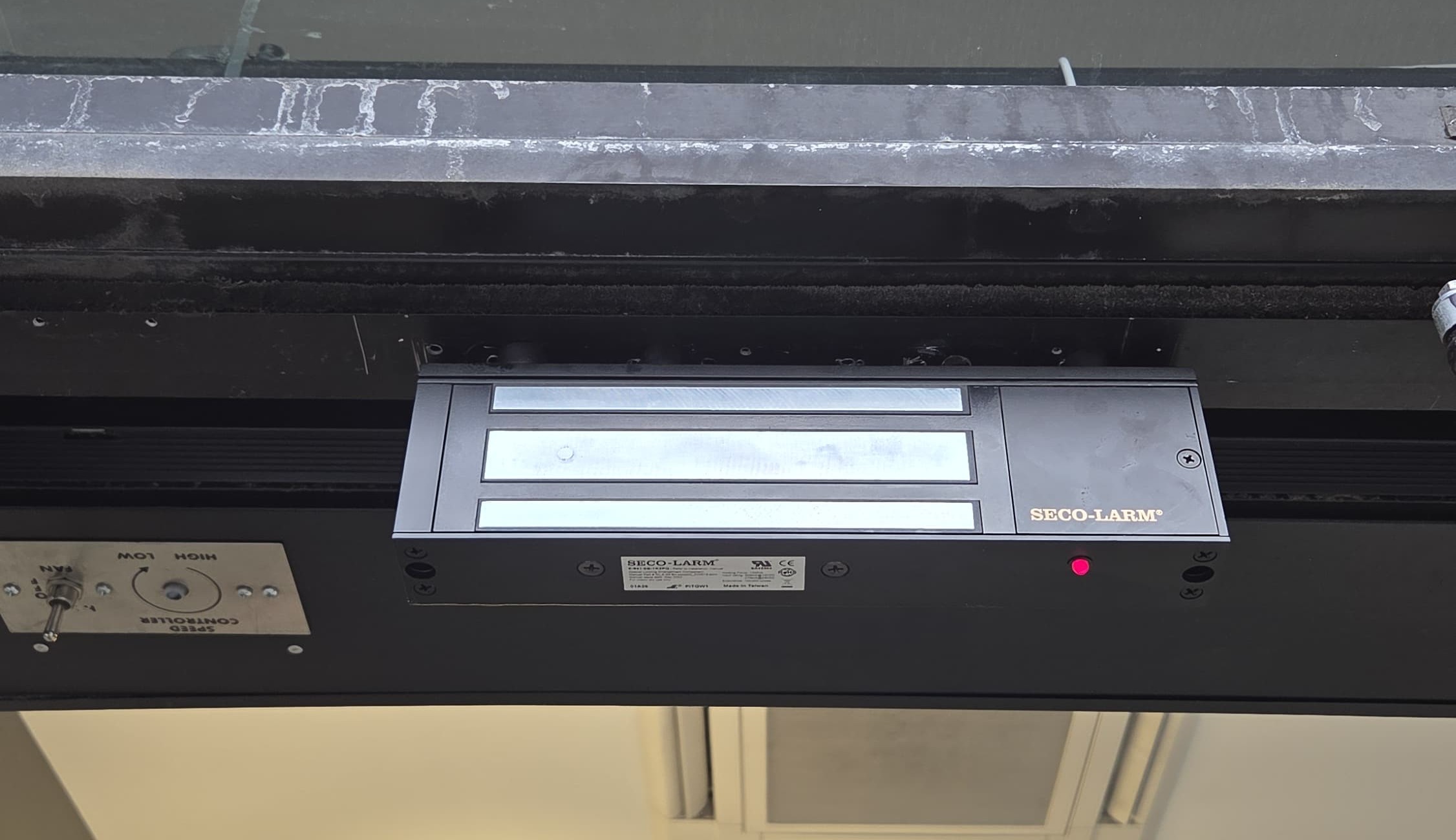 SECO-LARM electromagnetic lock (maglock) mounted on the header of a commercial glass entrance door, Bronx, NYC